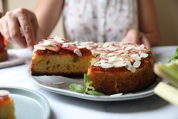 Woman taking piece of freshly baked rhubarb pie with almond flakes at white marble table, closeup