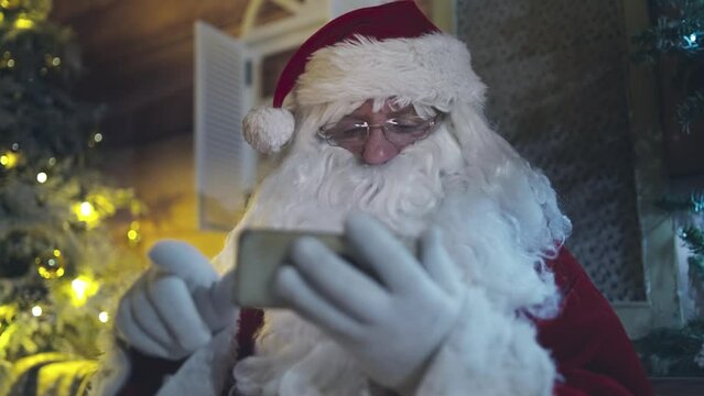 Santa Claus is sitting in a chair in front of the fireplace at night and using smartphone, scrolling and surfing on the internet. Christmas as a holiday concept.