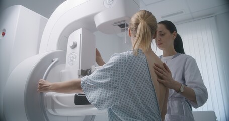 Hospital radiology room. Caucasian woman stands during mammography screening examination in clinic. Female doctor adjusts modern mammogram machine for patient, uses computer. Breast cancer prevention.