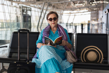 Tourist senior woman sitting on the bench in airport hall in waiting for her flight reading a book