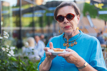 Senior woman in blue clothes and sunglasses holding a small plate with cake outside in summer
