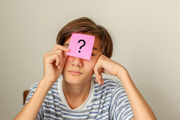 Thinking teenager boy, school student with question mark on paper on his forehead isolated on gray wall background. Confusing, doubt teenage concept. - obrazy, fototapety, plakaty