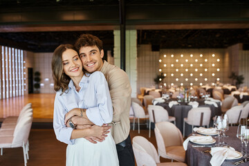 overjoyed man embracing girlfriend in banquet hall with decorated festive tables, wedding setup