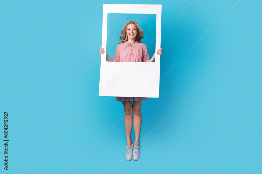 Wall mural Full size photo of lovely positive girl dressed stylish pink clothes jumping holding frame in hands isolated on blue color background