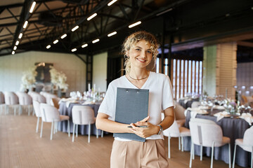 positive decorator with clipboard looking at camera near tables with festive setting in banquet hall