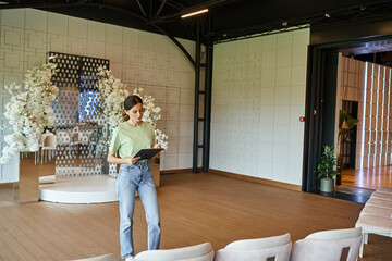 young event manager writing on clipboard in modern banquet hall with armchairs and floral decor