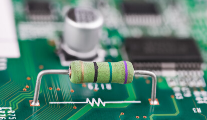 Closeup of resistor and white electronic symbol on green PCB surface. Small component with color code and two metal wire terminals on printed circuit board and blurry capacitor or chips in background.