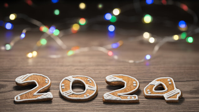 Close-up of gingerbread numerals on wood with colored LED light strings in blurry background with bokeh. Golden baked sweets for happiness in 2024 New Year. Christmas cookies decorated by white icing.