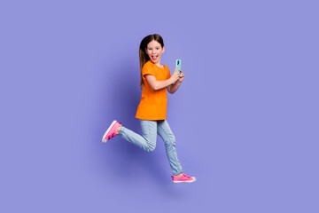 Fototapeta na wymiar Full size photo of positive schoolgirl wear stylish t-shirt hold smartphone run in empty space isolated on violet color background