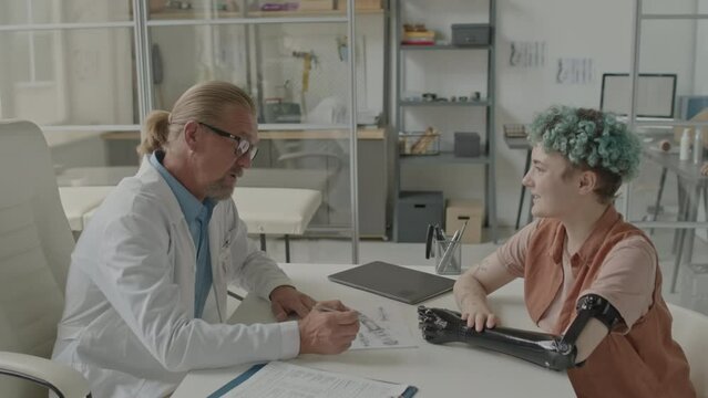 Waist up of Caucasian mid aged male prosthetist and young woman with bionic arm having conversation in modern doctors office in clinic