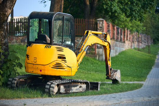 Tracked orange mini excavator digger near road with green trees and stone fence on background with copyspace.