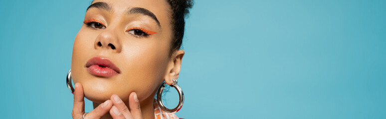 beautiful african american model in silver hoop earrings and rings looking at camera on blue, banner