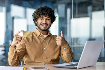 Portrait of a young Indian man, businessman, bank employee sitting in the office at a table with a laptop, holding a credit card in his hand and pointing super finger at the camera.