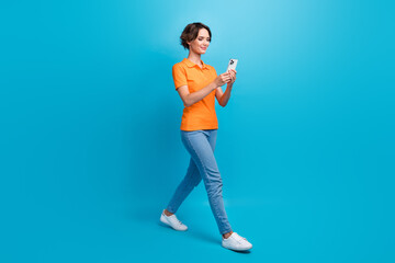 Fototapeta na wymiar Full length photo of nice woman wear orange t-shirt showing v-sign look at smartphone go empty space isolated on blue color background