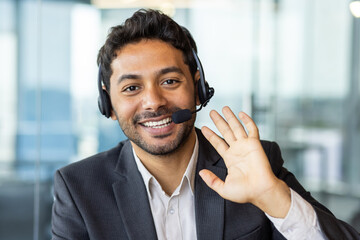 Close-up portrait of a young smiling Indian man in a suit sitting in the office, talking and waving...