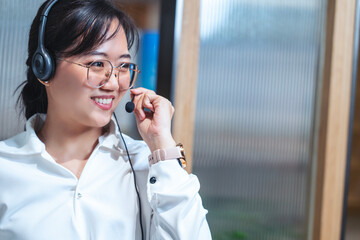 Asian woman in glasses, headphones and microphone working in console, answering telephone calls...