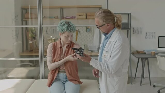 Medium shot of Caucasian mature prosthetics specialist fitting modern arm prosthesis to young female patient during medical consultation in contemporary clinic