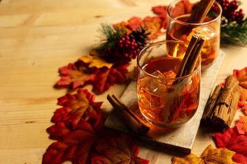 Top view of two glasses of whiskey with ice cubes and cinnamon sticks on a wooden table, with mapple leaves and black background
