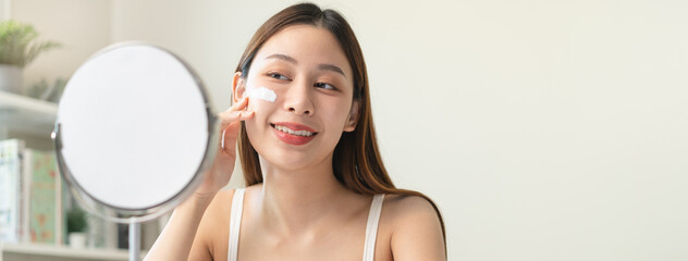 wellness care concept,  young woman looking at the mirror and applying facial skin care cream on her face