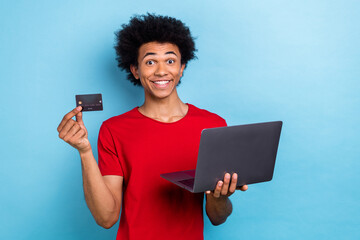 Photo of cheerful pleasant guy with afro hairstyle dressed red stylish t-shirt hold laptop credit card isolated on blue color background