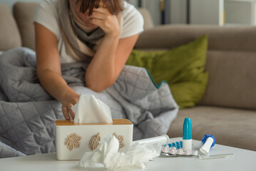 Medicines for colds and paper napkins are on the table, a woman with signs of flu and colds is sitting in the background and takes a napkin out of the box.
