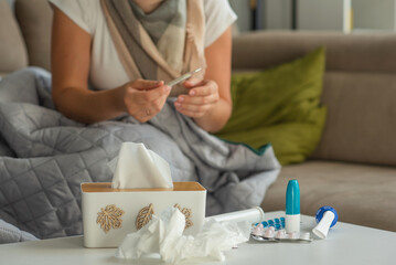 Medicines against colds and paper napkins are on the table, a woman with signs of flu and colds is sitting in the background and holding a thermometer in her hands.