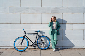 Young empowered ginger-haired woman in green suit talking on smartphone standing with bicycle at...