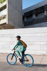 Stylish business woman in green suit, black helmet and yellow backpack riding her bicycle at the financial district with modern building on the background