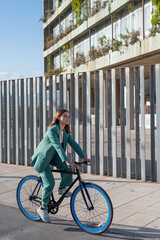 Young empowered ginger-haired woman in green suit riding a fixie at the financial district with modern building on the background