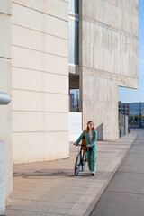 Young empowered ginger-haired woman in green suit walking with bicycle at the financial district with modern building on the background