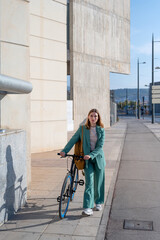 Young empowered ginger-haired woman in green suit walking with bicycle at the financial district with modern building on the background