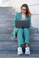 Business woman sitting on stairs outside office building while working with laptop