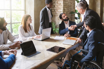Collaborative Business Handshake in Diverse Office: Young bearded man and senior silver-haired individual seal a deal amid diverse colleagues, including an inclusive member in a wheelchair.