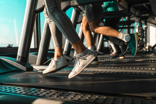 Picture of people wearing sports shoes running on treadmill in gym. Photo of people practicing cardio with only legs and feet in the frame.