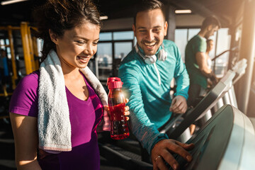 Happy athletic couple communicating while jogging on treadmills in a health club. Man explaining a woman how to turn on cardio machine. Attractive woman with water bottle needing assistance at gym. - obrazy, fototapety, plakaty