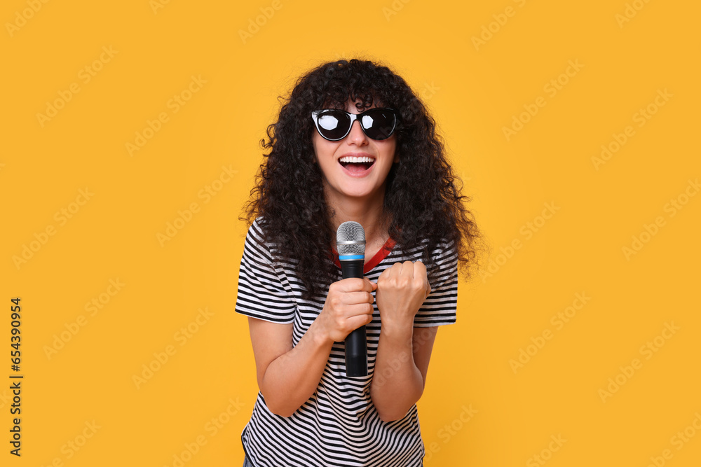 Poster Beautiful young woman with microphone and sunglasses singing on yellow background