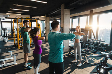 Group of athletic people exercising in a health club. Attractive female fitness instructor showing warming up exercises to her client in modern gym with city view.