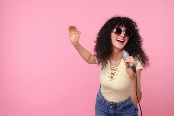 Beautiful young woman with microphone and sunglasses singing on pink background. Space for text