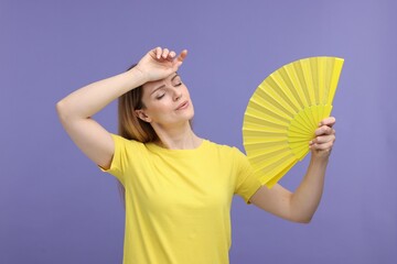 Beautiful woman waving yellow hand fan to cool herself on violet background