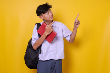 Young Asian student in school uniform looking above and pointing up to empty space aside for...