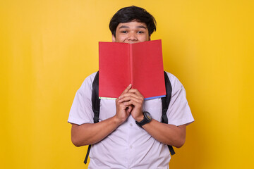 Funny young Asian student hiding his face behind book while standing over yellow background