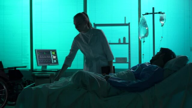 Full-sized silhouette video of a double amputee laying in a hospital bed. A female doctor, nurse comes in for a check-up. She studies his medical card and talks to a patient.