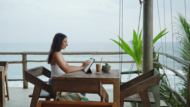 Young illustrator drawing on tablet at cafe with ocean view, palm trees. Pretty focused woman works remotely at authentic bar near tropical coast. Freelancer editing illustration at the seaside.