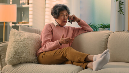 Happy old woman relaxing on cozy sofa talking on smartphone smiling senior grandma at comfy couch...
