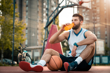 Tired basketball player drinking water from bottle while sitting at basketball court outdoors....