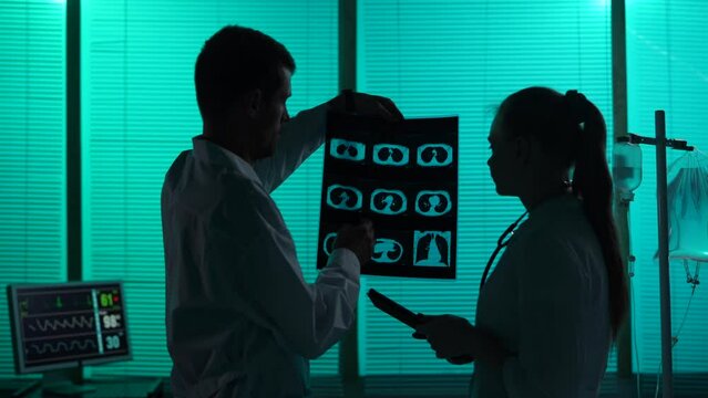 Medium-sized silhouette video of two doctors standing in a hospital ward. Doctors look at patient's lung ct, discussing the diagnosis and a treatment method.