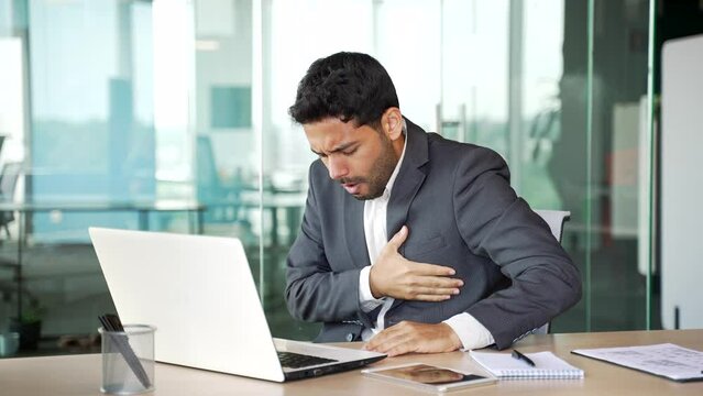 A young businessman in a suit has a heart attack working on a laptop sitting at a workplace in a business office. Sick entrepreneur suffers from tension in chest and holds the sore spot with his hand