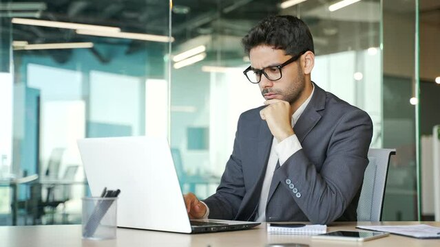 Serious thoughtful businessman in a formal suit works on a laptop while sitting at a workplace in a business office. Handsome entrepreneur in glasses is busy with a startup project on the computer