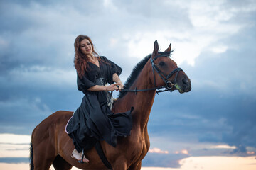 Obraz premium A beautiful young horsewoman in a black dress and with her hair down, riding a horse, portrait against the background of the evening sky, horseback riding in the open air