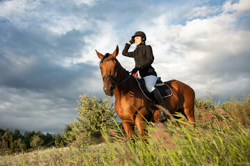 Horsewoman in equestrian sports gear, riding a horse, against an expressive sky, horseback riding in the open air - 654451183